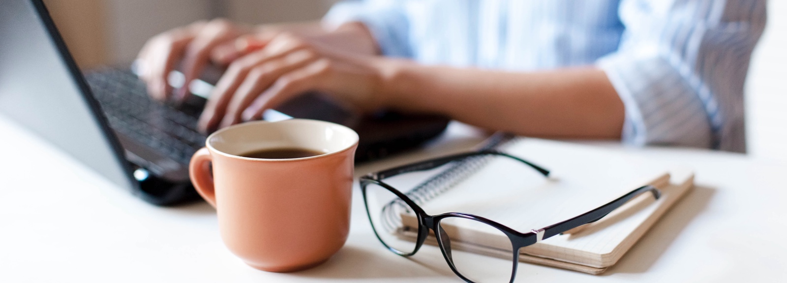 Close up of a person's hands on a laptop keyboard plus a coffee mug and glasses